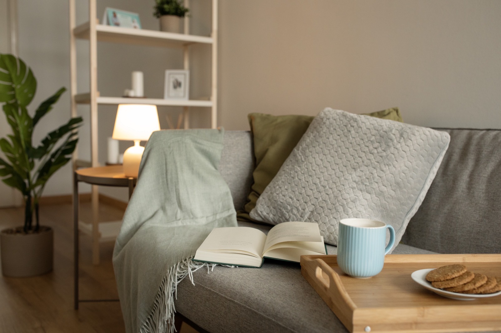 Un sofá con una manta, un libro abierto y una bandeja con una taza y galletas, en un salón iluminado por una lámpara
